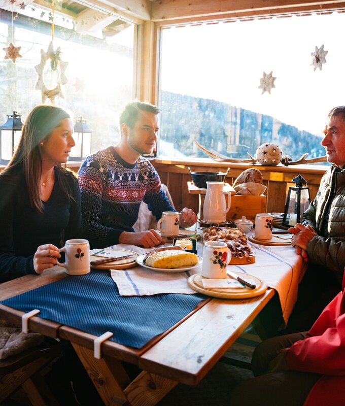Eine Gruppe von Personen genießt eine gemütliche Mahlzeit in einem rustikalen, winterlichen Innenraum. Der Blick schweift durch die großen Fenster auf eine schneebedeckte Landschaft. | © Urlaub am Bauernhof Kärnten / Daniel Gollner