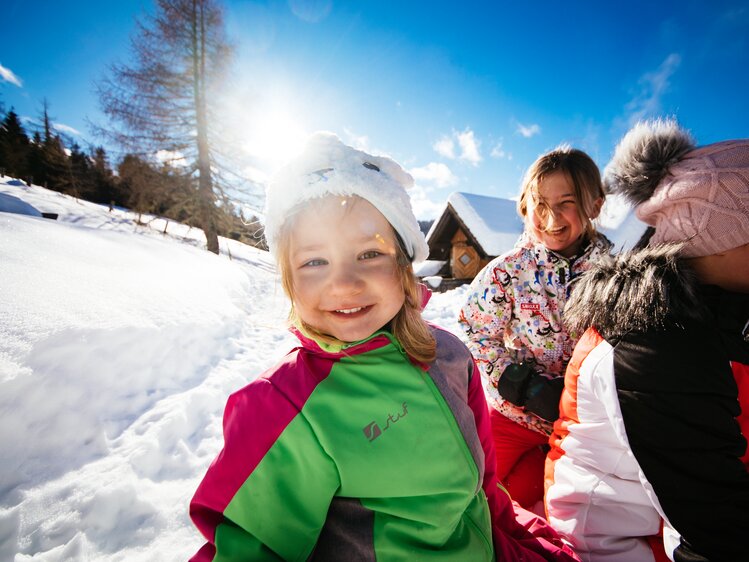 Glückliche Kinder, die im Winter beim Schneefall Spaß haben. Sie tragen warme Kleidung, Schal und Mütze, und lächeln in die Kamera. Im Hintergrund sind verschneite Berge und Häuser zu erkennen. | © Urlaub am Bauernhof Kärnten / Daniel Gollner