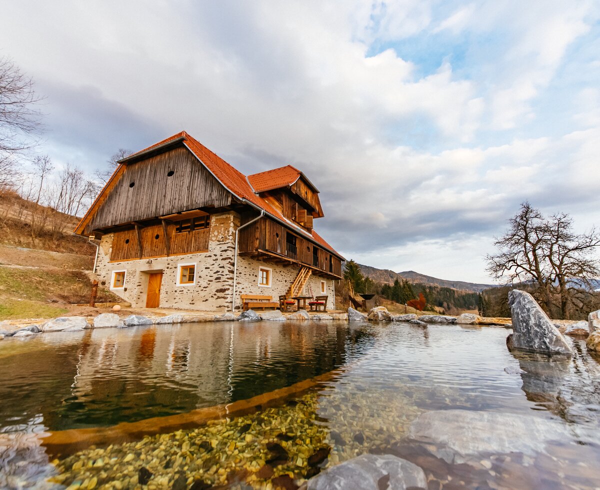 Ein malerisches Gebirgshaus mit Holzfassade und Strohdach direkt am Wasser, umgeben von Bäumen und Felsen. Ein ruhiger See spiegelt die Gebäude und die Wolken am Himmel wider. Die Szene strahlt Natürlichkeit und Ruhe aus. | © Urlaub am Bauernhof Kärnten / Daniel Gollner