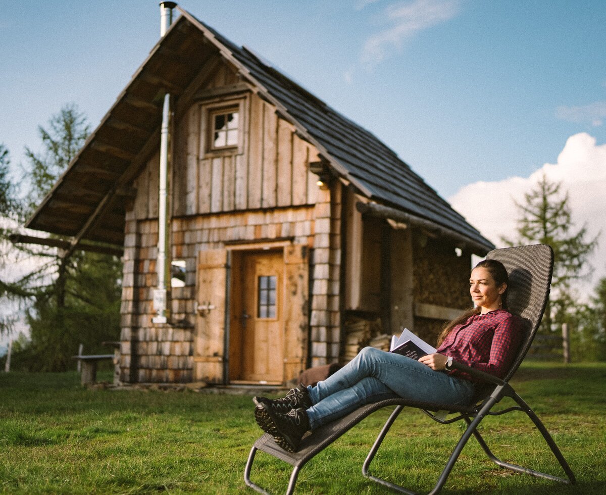 Eine Person sitzt in einem Campingstuhl vor einer alten, holzverkleideten Hütte in einer ländlichen Umgebung und genießt die Natur, während sie ein Buch liest. | © Urlaub am Bauernhof / Daniel Gollner