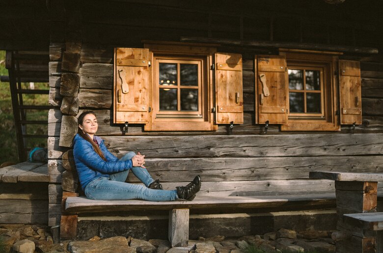 Eine Frau in blauer Jacke sitzt auf einer Holzbank vor einer rustikalen Hütte mit Fensterläden. Die Hütte ist von Holz und befindet sich in einer natürlichen Umgebung. | © Urlaub am Bauernhof / Daniel Gollner