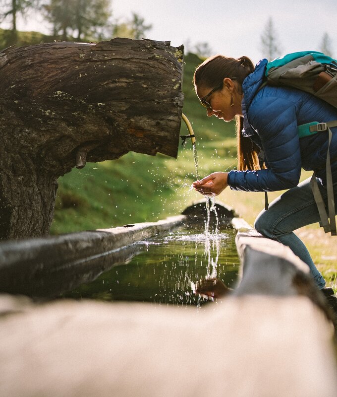 Wanderin hockt an einem Waldbach und trinkt aus einem Holzstamm, der als Wasserkrug dient. Die natürliche Umgebung mit Bäumen und Wiesen schafft eine friedliche Stimmung. | © Urlaub am Bauernhof / Daniel Gollner