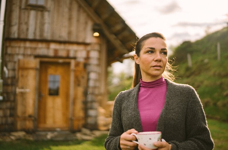 Eine Frau trinkt Tee vor der Almhütte. | © Urlaub am Bauernhof / Daniel Gollner Eine Frau in einem grauen Pullover und einer rosa Jacke steht vor einer alten Holzhütte auf einer Wiese und hält eine Tasse in der Hand. Windspiele treiben in der Luft, was eine ruhige Atmosphäre vermittelt. | © Urlaub am Bauernhof / Daniel Gollner