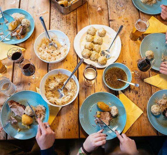 Gut gefüllte Tische mit Tellern voller hausgemachter Speisen, wie Knödel, Eintöpfe und Bier. Die Hände der Gäste greifen nach den Utensilien, um das Essen zu genießen. Eine gesellige Atmosphäre in einem gemütlichen, hölzernen Ambiente. | © Urlaub am Bauernhof / Daniel Gollner