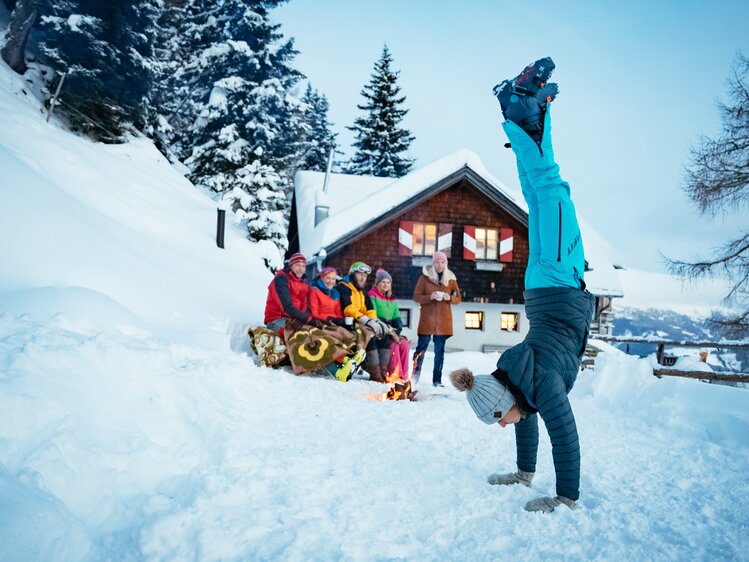 Gruppe von Wintersportlern vor einer gemütlichen Hütte im verschneiten Wald, eine Person macht einen Handstand im Schnee. | © Urlaub am Bauernhof Kärnten/ Daniel Gollner