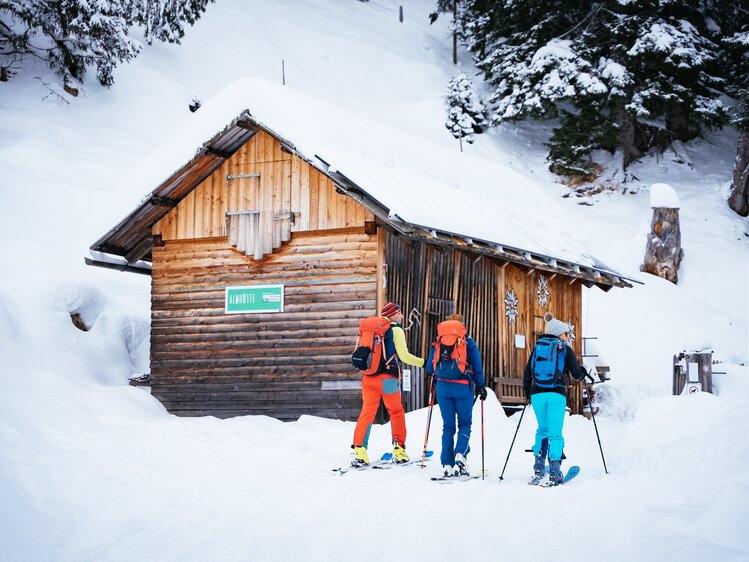 Eine Holzhütte inmitten verschneiter Wälder, vor der drei Skifahrer in bunter Winterkleidung stehen. Die Szene vermittelt Entspannung und den Genuss der winterlichen Natur. | © Urlaub am Bauernhof Kärnten/ Daniel Gollner