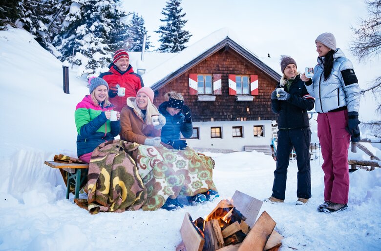 Gruppe von Freunden, die bei einem verschneiten Berghüttchen eine gemütliche Winterrast machen. Sie sitzen um ein Lagerfeuer herum und genießen heiße Getränke. Die Hütte im traditionellen Holzbau-Stil liegt inmitten einer idyllischen, winterlichen Landschaft. | © Urlaub am Bauernhof Kärnten/ Daniel Gollner