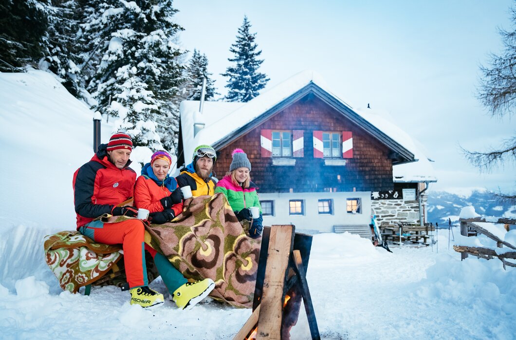 Vier Winterurlauber sitzen beschützt vor der Kälte am Feuer vor einer verschneiten, rustikalen Holzhütte. | © Urlaub am Bauernhof Kärnten/ Daniel Gollner