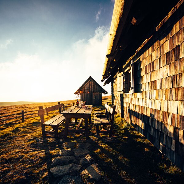 In der Ferne steht eine hölzerne Hütte mit Schindeldach, umgeben von einer Bank und Picknick-Tischen. Der Vordergrund zeigt eine sonnenbeschienene Landschaft mit Zäunen und hohem Gras. | © Urlaub am Bauernhof Kärnten/ Daniel Gollner 