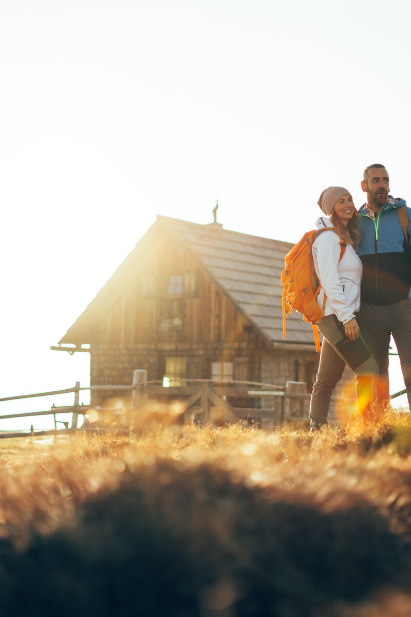 Paar in moderner Wanderkleidung steht vor reetgedecktem Haus, Landschaft im Sonnenlicht, Hügel und Zaun im Vordergrund.
