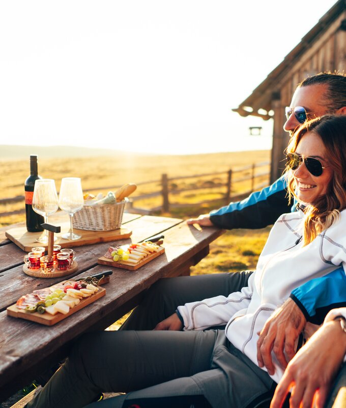Zwei Freunde genießen einen Picknick-Nachmittag in einer ländlichen Umgebung. Vor ihnen steht ein Tisch mit Wein, Käse und Obst. Sie sitzen entspannt und blicken in die Ferne.