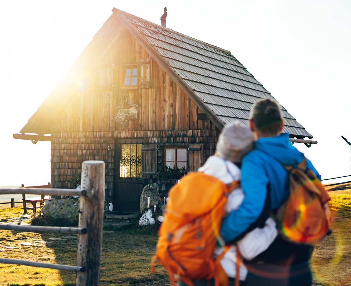 Traditionelles Holzhaus mit Reetdach, von Wanderern mit Rucksäcken besucht, inmitten einer Landschaft mit Zaun und Baum. Warmes, sonniges Licht umspielt die Szenerie.