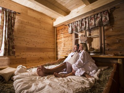 Paar im Heubett | © Martin Steinthaler / Urlaub am Bauernhof Kärnten Ein gemütliches Holzhütten-Interieur mit einem Bett aus Naturmaterialien, auf dem eine verliebtes Paar entspannt beisammensitzt, umgeben von Dekorationen im rustikalen Stil. | © Martin Steinthaler / Urlaub am Bauernhof Kärnten