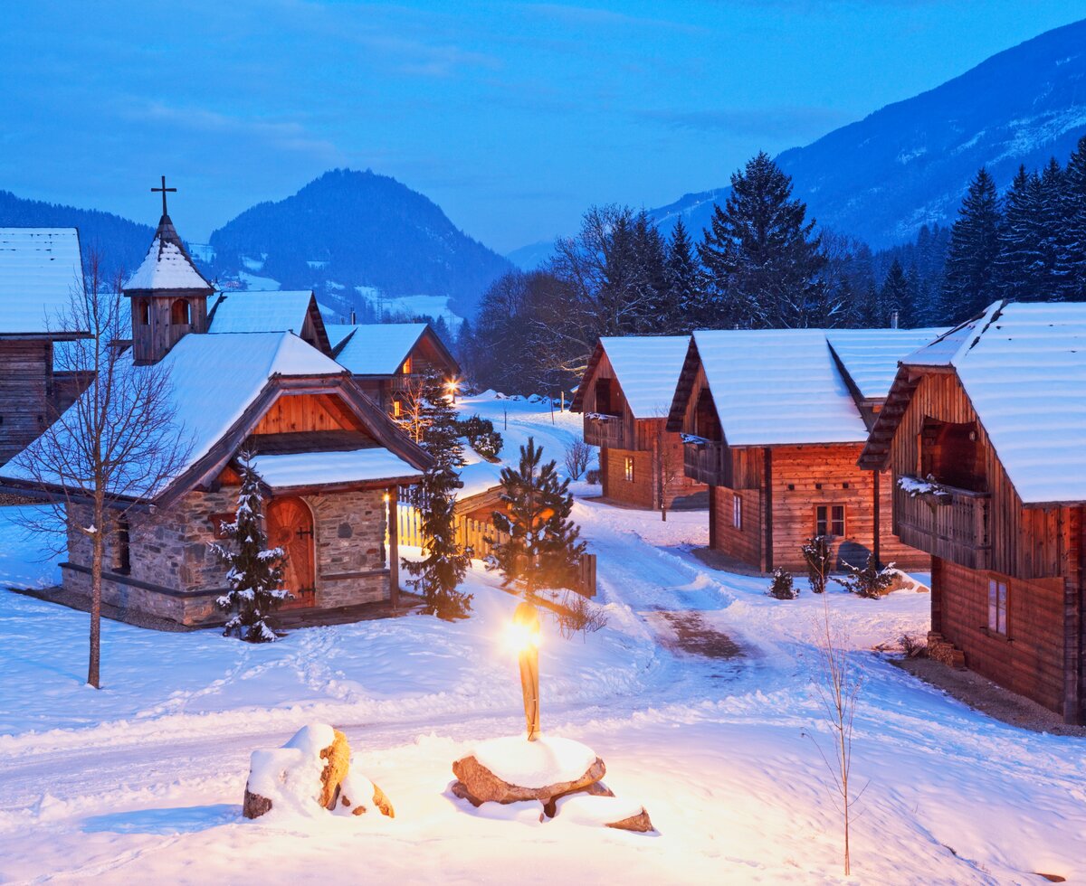 Eine verschneite Bergidylle mit malerischen Holzhäusern, einer Kirche und beleuchteten Weihnachtstannen. Eine abendliche Winterstimmung mit sanftem Lichtschein in der verschneiten Landschaft. | © Helene Hartweger Glanzer / Landgut Moserhof
