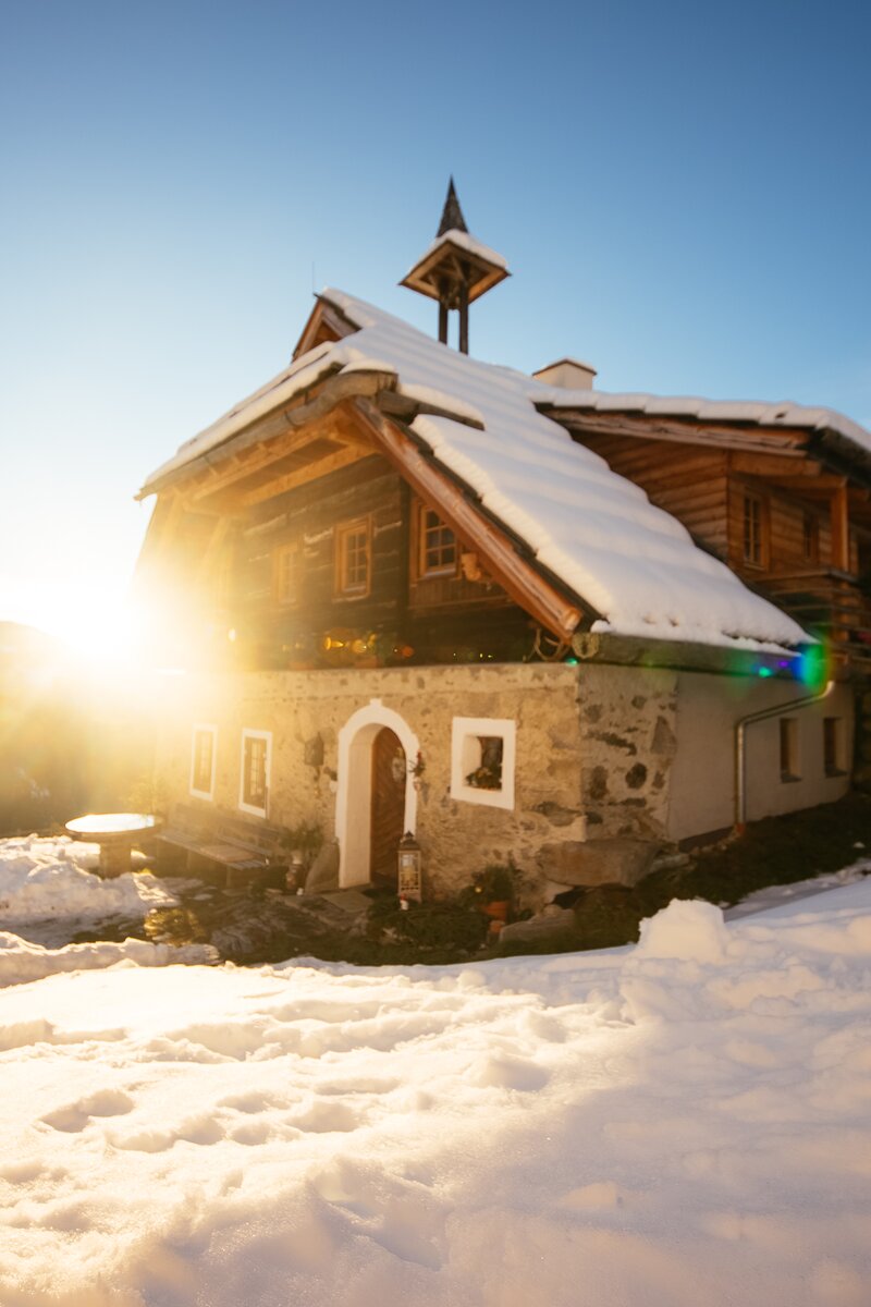 Altes, traditionelles Holzhaus mit Satteldach in verschneiter, bergiger Landschaft. Sonniges Licht erfüllt die Szene, verleiht dem Haus eine verträumte, idyllische Stimmung. | © Urlaub am Bauernhof Kärnten / Daniel Gollner