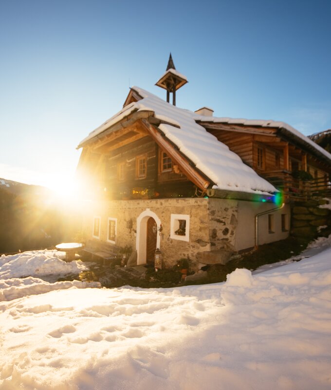 A traditional alpine cabin nestled in a snowy landscape, illuminated by the warm glow of the setting sun. | © Urlaub am Bauernhof Kärnten / Daniel Gollner