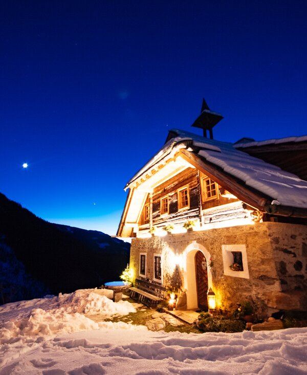 Holzhütte in verschneiter Berglandschaft bei Nacht, beleuchtet durch Fackelschein und Sterne am tiefblauen Nachthimmel. | © Daniel Gollner / Urlaub am Bauernhof Kärnten 