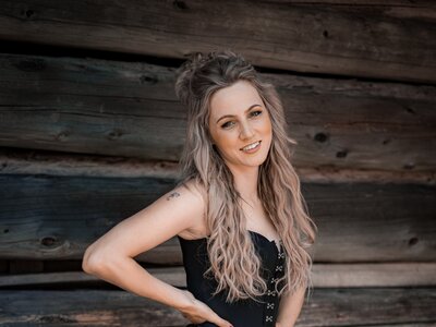 Melissa Naschenweng | © Lambauer Entertainment / Christoph Hatheuer Junge Frau mit langen, blonden Haaren und Fringerock in Schwarztönen und leuchtendrosa. Sie steht lächelnd vor einer Holzwand. | © Lambauer Entertainment / Christoph Hatheuer