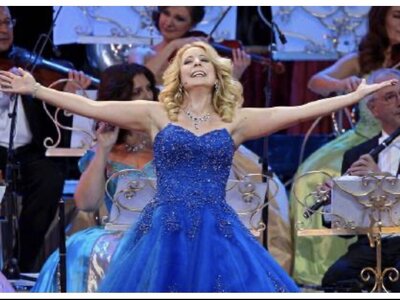 Sängerin Iva Schell | © Urlaub am Bauernhof / Schell Eine Frau im prachtvollen blauen Ballkleid mit ausgebreiteten Armen und strahlendem Lächeln, umgeben von einer Orchesterkulisse. | © Urlaub am Bauernhof / Schell