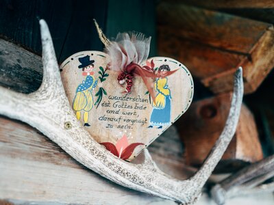 Herz aus Holz | © Urlaub am Bauernhof Kärnten / Daniel Gollner Ein bemaltes Herz an einem verwitterten Holzbrett, geschmückt mit Federn und einer bunten Blume. Darauf befindet sich eine handschriftliche Aufschrift in deutscher Sprache. | © Urlaub am Bauernhof Kärnten / Daniel Gollner