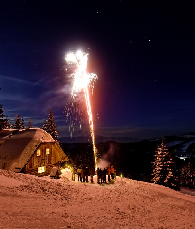 Silvester auf der Alm  | © Urlaub am Bauernhof/ Tom Lamm Eine Menschengruppe versammelt sich vor einer beleuchteten Hütte, während oben am Nachthimmel bunte Feuerwerksexplosionen zu sehen sind. Die Szene vermittelt eine winterliche Feststimmung in einer bergigen Landschaft. | © Urlaub am Bauernhof/ Tom Lamm