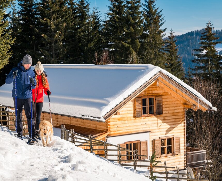 Ein hölzernes Blockhaus umgeben von verschneiter Berglandschaft. Ein Paar in Winterkleidung und ein Hund bewegen sich auf einem Pfad durch den Schnee. Die Sonne scheint auf das Haus und den Winterwald. | © Michael Stabentheiner