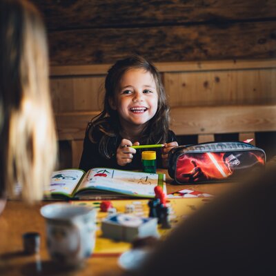 Ein junges, lächelndes Mädchen sitzt an einem Tisch in einem Holzhaus und malt in einem Malblock. Auf dem Tisch liegen verschiedene Mal- und Schreibutensilien wie Stifte, Lineal und Bücher verstreut. | © Urlaub am Bauernhof Salzburg / Daniel Gollner 