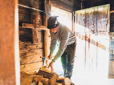 Beim Holzhacken auf der Alm | © Urlaub am Bauernhof Salzburg / Daniel Gollner Man in workwear chopping firewood in a rustic wooden cabin interior, with shelves and art on the walls. | © Urlaub am Bauernhof Salzburg / Daniel Gollner