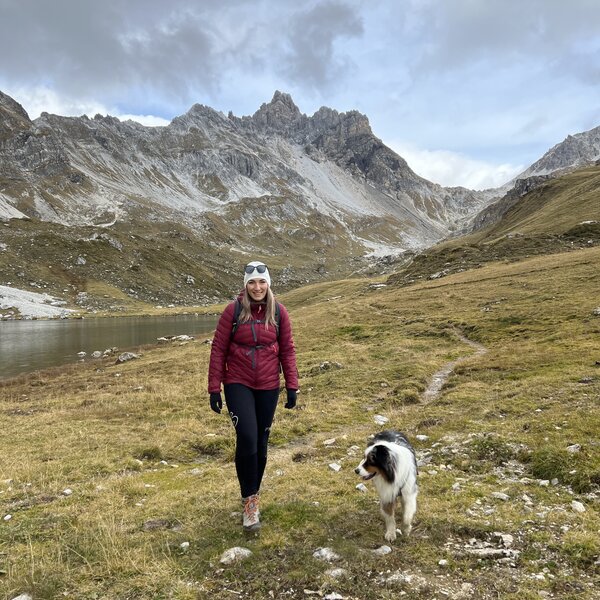A woman in a red jacket walking with a dog in a mountainous, grassy landscape with snow-capped peaks in the background. | © Urlaub auf der Alm / Julia Winkler