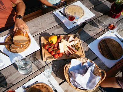 Almjausen auf einer Hütte in der Steiermark | © Urlaub am Bauernhof Steiermark / Daniel Gollner Rustic wooden table with fresh bread, cheese, tomatoes, and glasses of red wine. Outdoor setting with vibrant colors and textures. | © Urlaub am Bauernhof Steiermark / Daniel Gollner