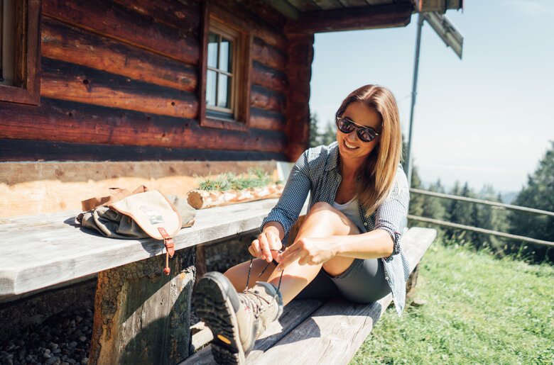 Eine junge Frau in Casual-Kleidung sitzt lächelnd auf einer Holzbank vor einer Berghütte und bindet ihre Wanderstiefel. Sie trägt Sonnenbrille und genießt die Aussicht auf eine ländliche Umgebung. | © Urlaub am Bauernhof Steiermark / Daniel Gollner 