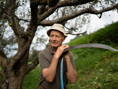 Altbauer Bacher hat immer eine Geschichte parat | © Urlaub am Bauernhof Kärnten / Daniel Gollner Mann lehnt auf der Sense | © Urlaub am Bauernhof Kärnten / Daniel Gollner