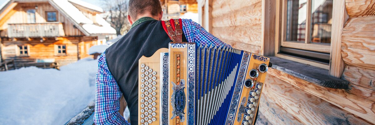 Musiker geht mit einer Steirischen Ziehharmonika am Rücken an einer Hütte vorbei | © Urlaub am Bauernhof Oberösterreich / Pascal Baronit