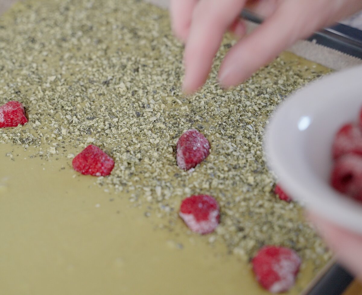Eine Hand füllt frische Himbeeren in eine Schüssel mit zerkleinerter grüner Masse, die möglicherweise zu Kräutern oder Müsli verarbeitet wird.