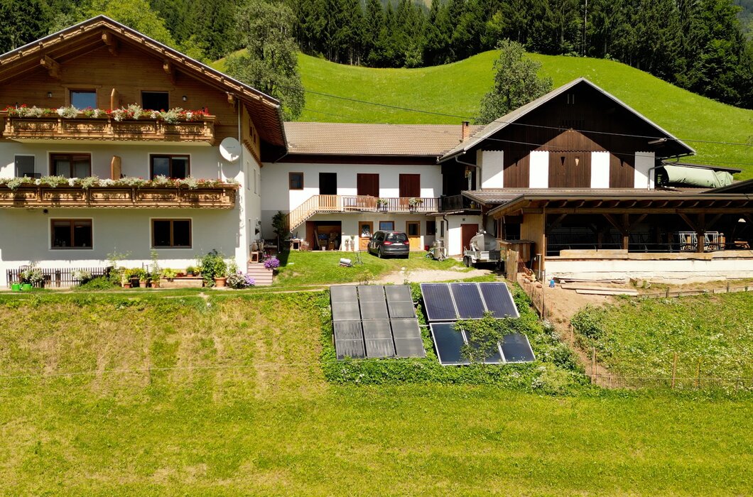 Ein großes, historisches Holzhaus mit Balkonen und Dachgauben, umgeben von grünen Wiesen und Wäldern. Vor dem Haus befinden sich Solarzellen zur Energiegewinnung. | © Urlaub am Bauernhof / Daniel Gollner