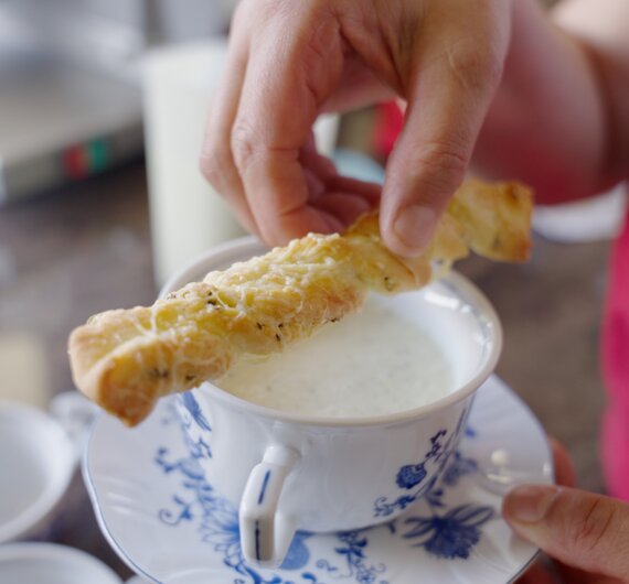 Eine Person in einem roten Pullover taucht ein knuspriges Brotstück in eine Tasse mit Flüssigkeit ein.