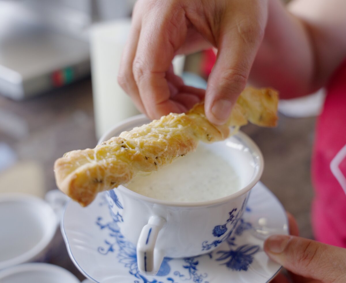 Eine Person in einem roten Pullover taucht ein knuspriges Brotstück in eine Tasse mit Flüssigkeit ein.