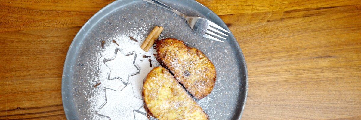 Auf einem grauen Teller liegen zwei goldbraune, mit Puderzucker bestäubte Gebäckstücke. Neben ihnen sind Sterne aus Puderzucker dekoriert. Vor dem Hintergrund einer Holzoberfläche lässt sich eine gabelähnliche Besteckposition erkennen. | © Urlaub am Bauernhof / Daniel Gollner