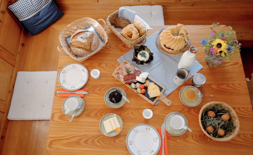 Einen gedeckten Holztisch mit verschiedenen Brotsorten, Käse, Marmelade, Obst, Blumengesteck und Getränken, eine herzhafte und gemütliche Frühstücksszene. | © Urlaub am Bauernhof / Daniel Gollner