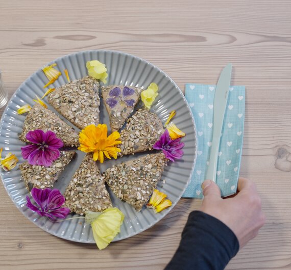 Teller mit verschiedenen Vollkornbrötchen, dekoriert mit bunten Blumen und Blättern, auf Holztisch mit blauer, gepunkteter Serviette. | © Urlaub am Bauernhof / Daniel Gollner
