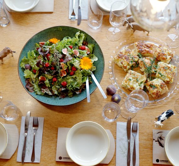 Ein gedeckter Esstisch mit verschiedenen Speisen, darunter ein grüner Salat und gebackene Gerichte, umgeben von Geschirr, Gläsern und Besteck, bereit für ein entspanntes Mittagessen oder Abendessen. | © Urlaub am Bauernhof / Daniel Gollner