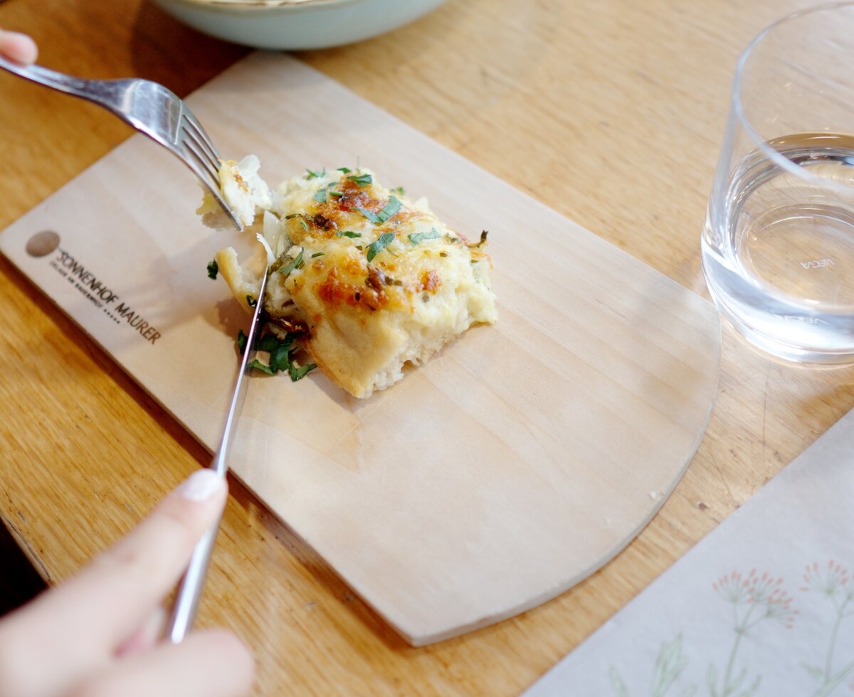 Ein Holzbrett mit einer Portion gegrillten Kartoffelauflaufs, dazu ein Glas Wasser auf einem Holztisch. | © Urlaub am Bauernhof / Daniel Gollner