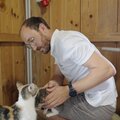 Ein Mann in weißem T-Shirt streichelt eine Katze in einer Holzhütte. Er trägt eine Brille und eine Armbanduhr und scheint die Katze liebevoll zu behandeln. | © Urlaub am Bauernhof / Daniel Gollner