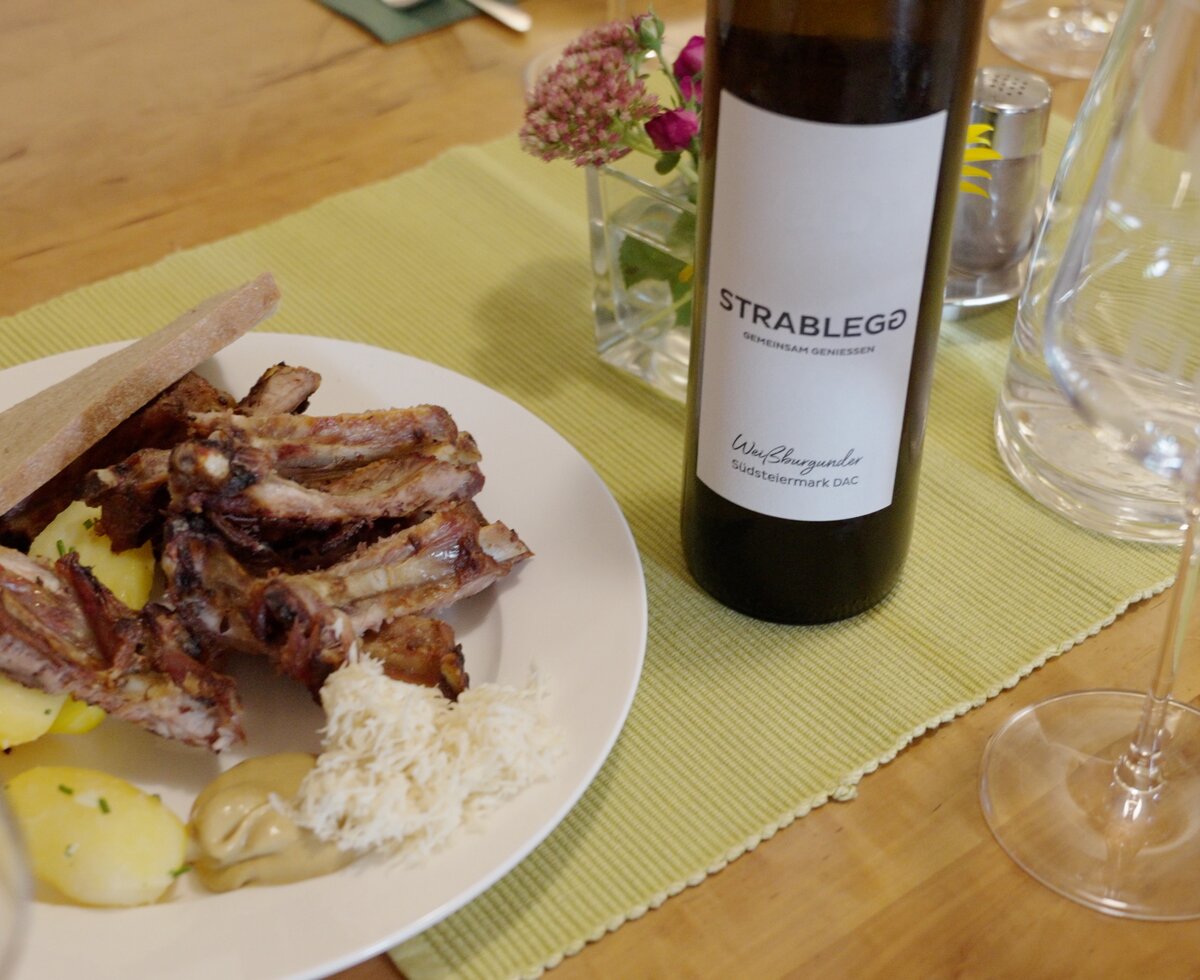 Auf dem Tisch befinden sich ein Teller mit gegrilltem Fleisch, Brot und Kartoffeln sowie eine Flasche Rotwein und Weingläser. Eine kleine Vase mit Blumen rundet die Tischeinstellung ab. | © Urlaub am Bauernhof / Daniel Gollner