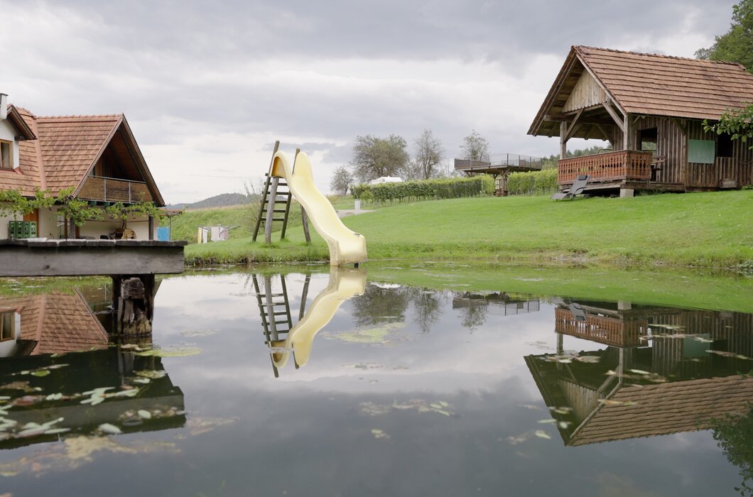 Ruhige, idyllische Landschaft mit Teich, Holzhäusern, Rutsche und grüner Wiese, die sich in der Wasseroberfläche widerspiegeln. | © Urlaub am Bauernhof / Daniel Gollner