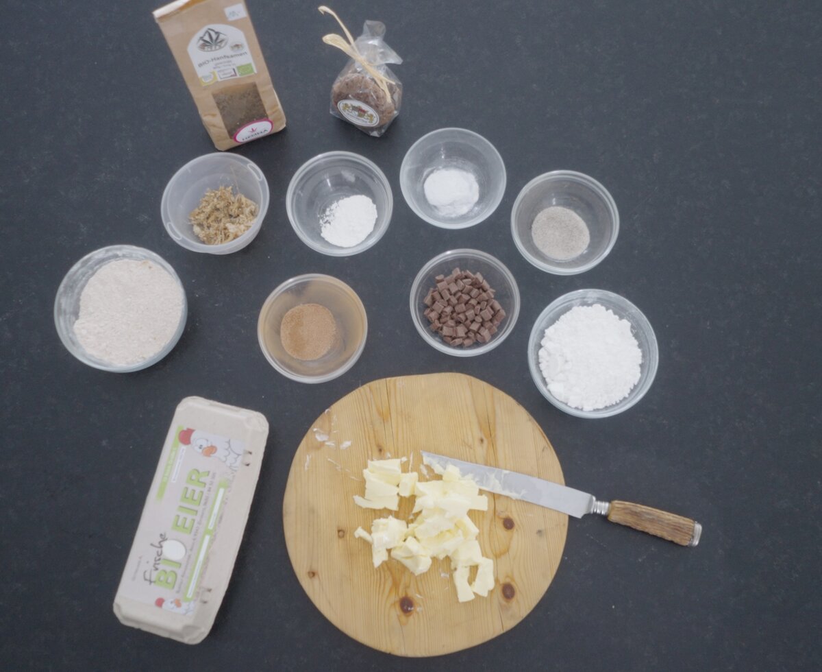 Verschiedene Zutaten auf einem Holzbrett, einschließlich Mehl, Zucker, Schokoladenchips und einer Schüssel, die für das Backen einer Speise oder den Verkauf vorbereitet sind. | © Urlaub am Bauernhof / Daniel Gollner