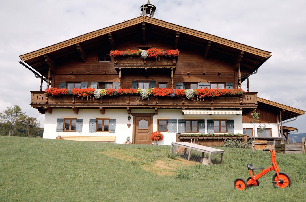 Typisches bayerisches Bauernhaus mit Holzfassade, geschmückten Fensterbänken und Blumenkästen, umgeben von einer Wiese und einem roten Kinderfahrrad. | © Urlaub am Bauernhof / Daniel Gollner