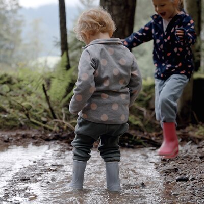 Two children walking on a muddy path in a forest, one wearing a gray polka dot jacket and the other in a navy blue patterned jacket, both wearing rain boots. | © Urlaub am Bauernhof / Daniel Gollner