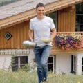 Holzhaus mit Blumenkästen und Balkon in üppiger Berglandschaft, davor Person in weißem Hemd und Jeans, Blick auf die Bergwelt. | © Urlaub am Bauernhof / Daniel Gollner