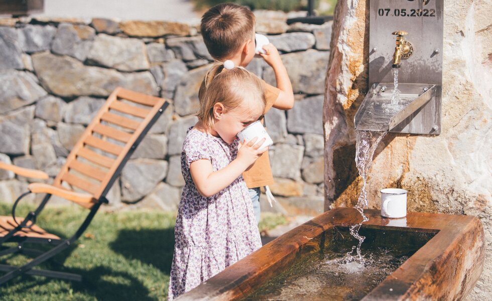 Zwei Kinder spielen an einer rustikalen Brunnenanlage in einer steinigen Umgebung. Der Junge scheint dem Mädchen zu helfen, sich zu waschen. Daneben steht ein Holzstuhl, der auf eine gemütliche Sitzecke hindeutet. | © Urlaub am Bauernhof / Daniel Gollner